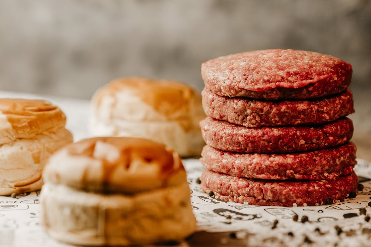 category-03 A stack of fresh raw beef patties with nearby burger buns, ready for cooking.